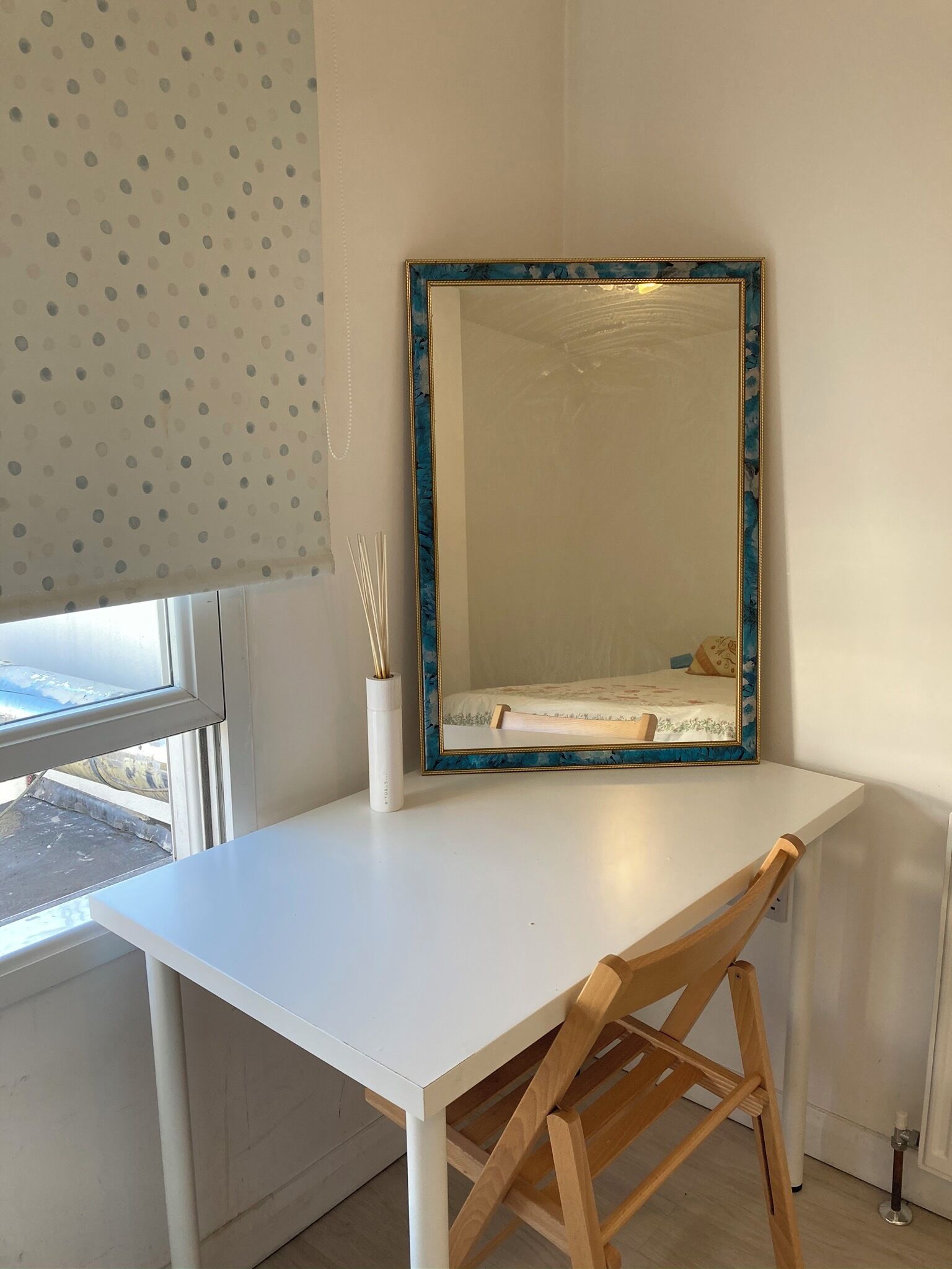 Un escritorio blanco con un espejo de marco azul, una silla de madera y un jarrón blanco con ramas de juncos.