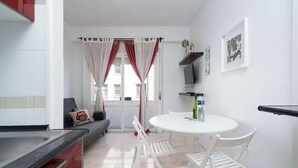 Ein kompakter Wohnraum mit Küchenzeile, einem Essbereich mit rundem Tisch und Stühlen, einer Couch mit Zierkissen und einem Fenster mit Vorhängen.
