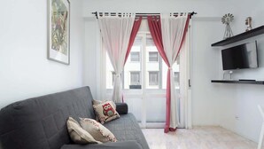 Ein modernes Wohnzimmer mit einem grauen Sofa, zwei Zierkissen, einem Flachbildfernseher auf einem schwarzen Standfuß und einem Fenster mit roten Vorhängen.