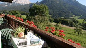 A balcony with a table set for two, a bottle of wine, and a menu. The balcony overlooks a mountainous landscape with green fields and red flowers.
