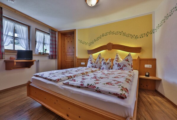 A bedroom with a wooden bed, a nightstand, a wall-mounted shelf, and a window with curtains.