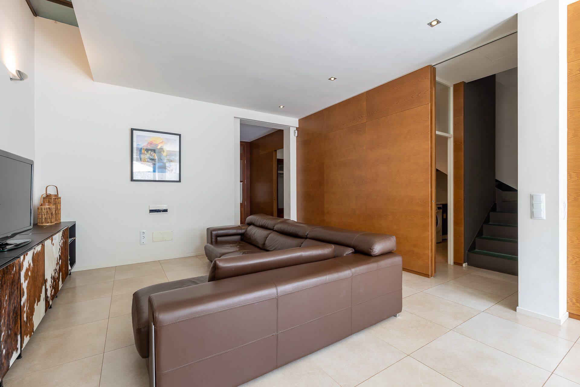 A modern living room with a large L-shaped brown leather sofa, a flat-screen TV, and a wooden cabinet.