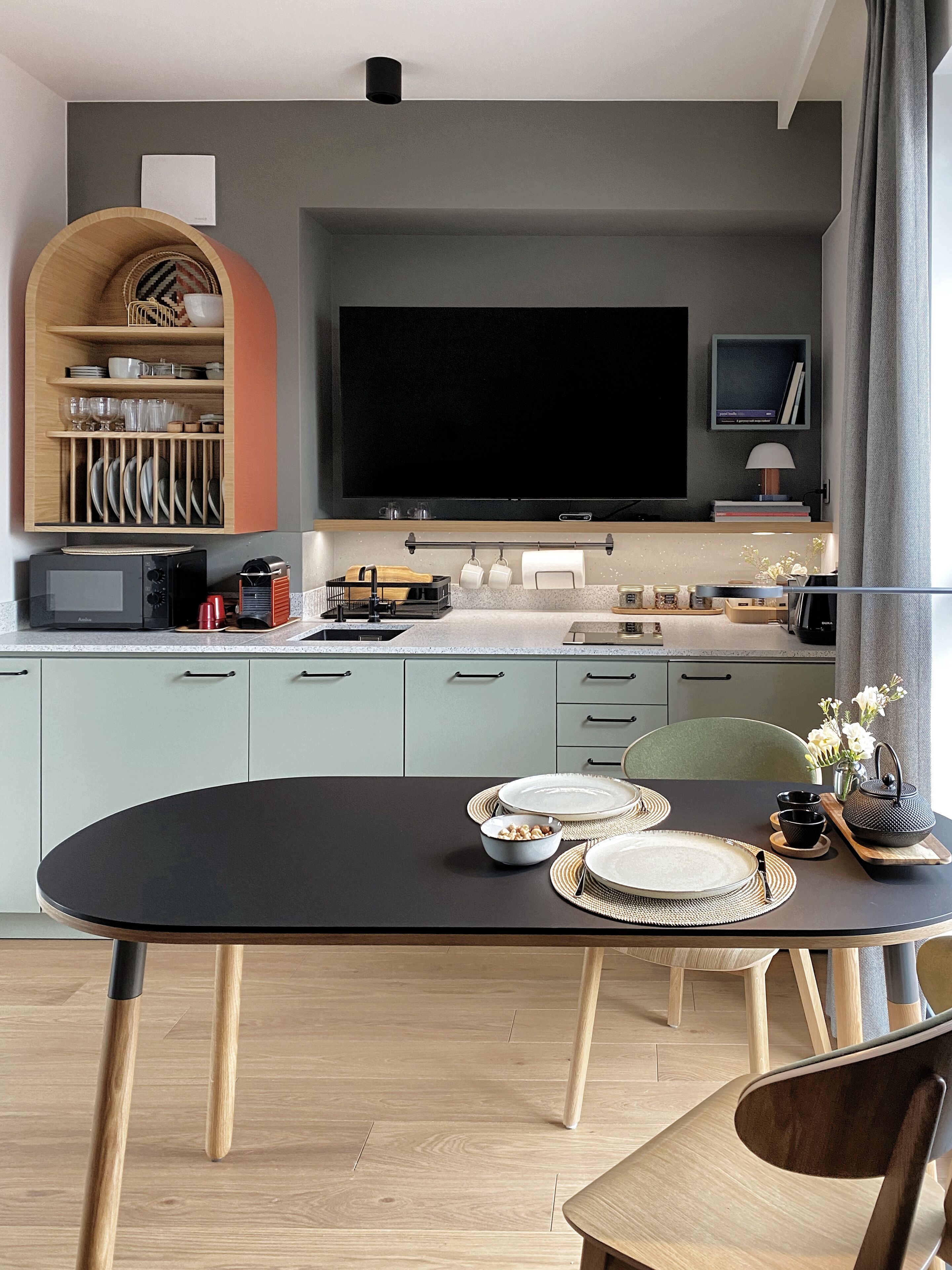 Una cocina moderna con una mesa de comedor para cuatro, un televisor de pantalla plana sobre la encimera y un armario de madera con vajilla.