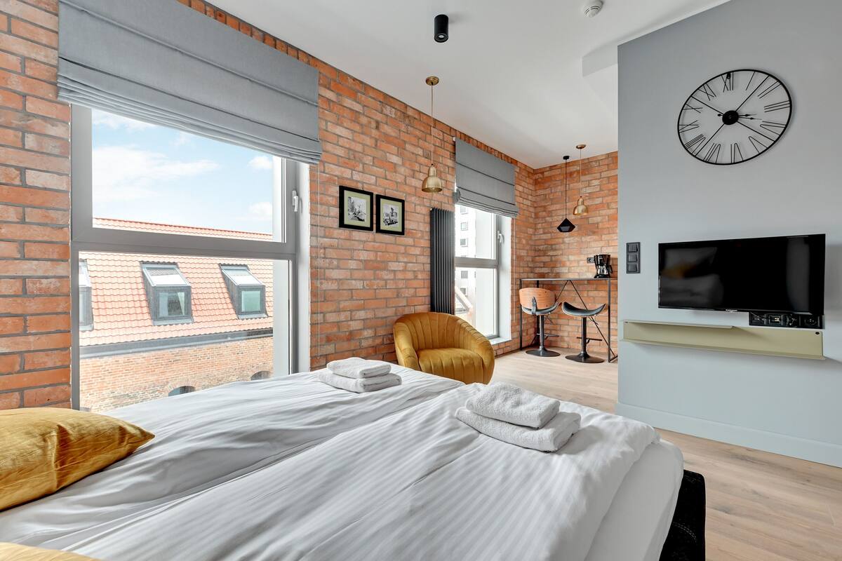 A modern hotel room with a brick wall, large windows, a bed with white linens, a mustard-colored armchair, a flat-screen TV, and a wall-mounted clock.