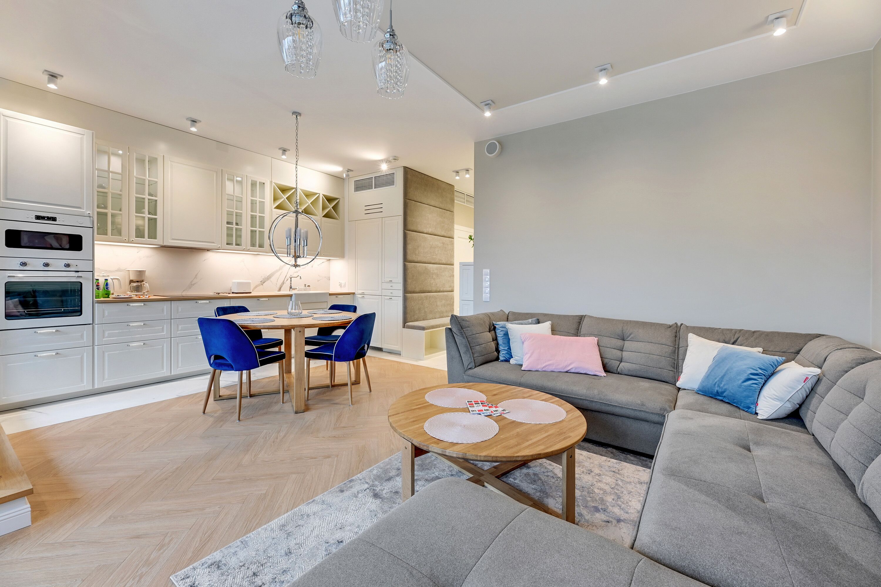 Uma sala de estar moderna com um sofá seccional cinza, uma mesa de centro de madeira e uma área de jantar com cadeiras azuis e uma mesa branca.