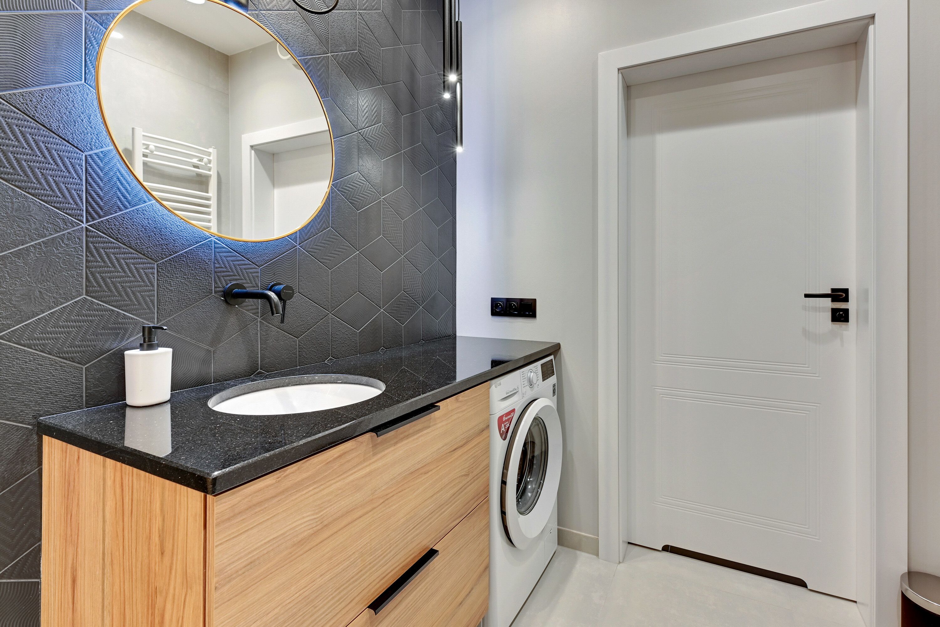 A modern bathroom with a black countertop, a white sink, a round mirror, and a washing machine.