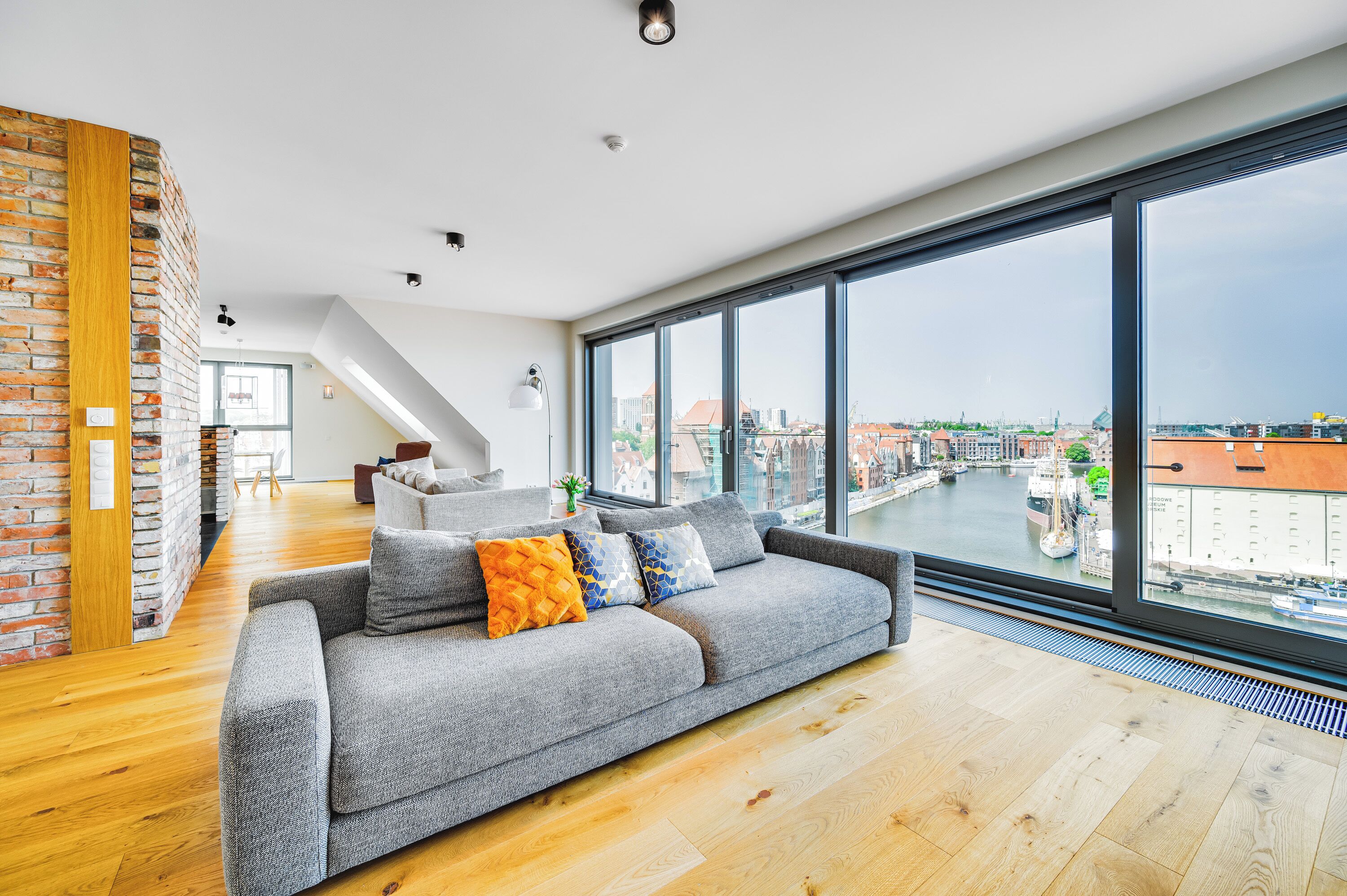 A modern living room with a grey sofa, wooden flooring, and large windows offering a view of a cityscape.