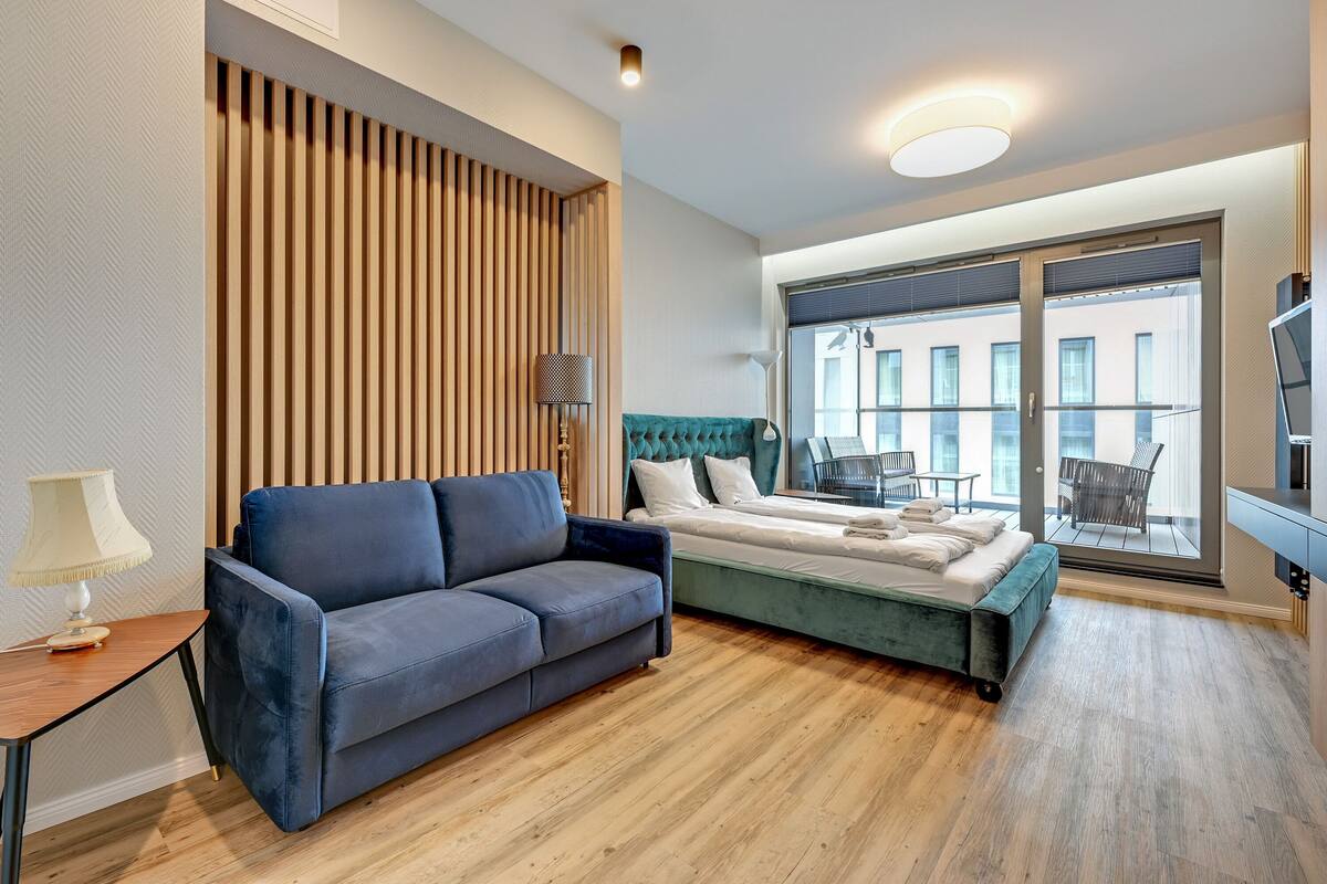 A modern hotel room with a blue sofa, a bed with a green headboard, a wooden slat wall, a large window with a view of buildings, and a TV on the right.