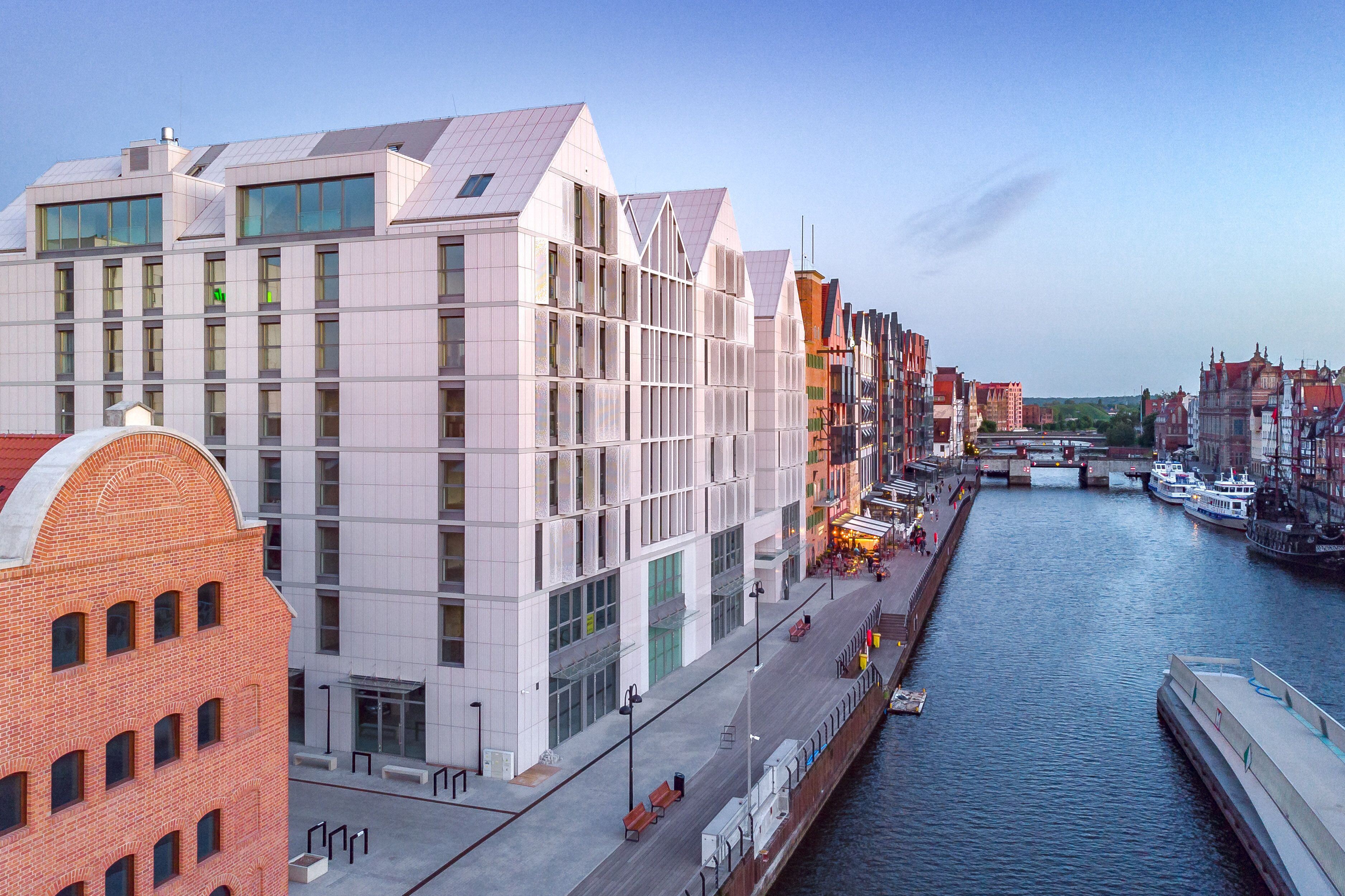 A modern building with a glass facade, a brick structure to the left, and a canal with boats.