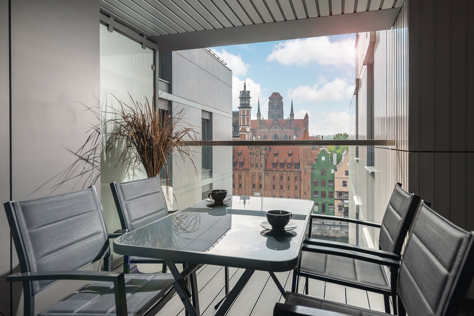 Un balcón con una mesa y sillas de vidrio, un florero con plantas secas y vistas a edificios históricos.
