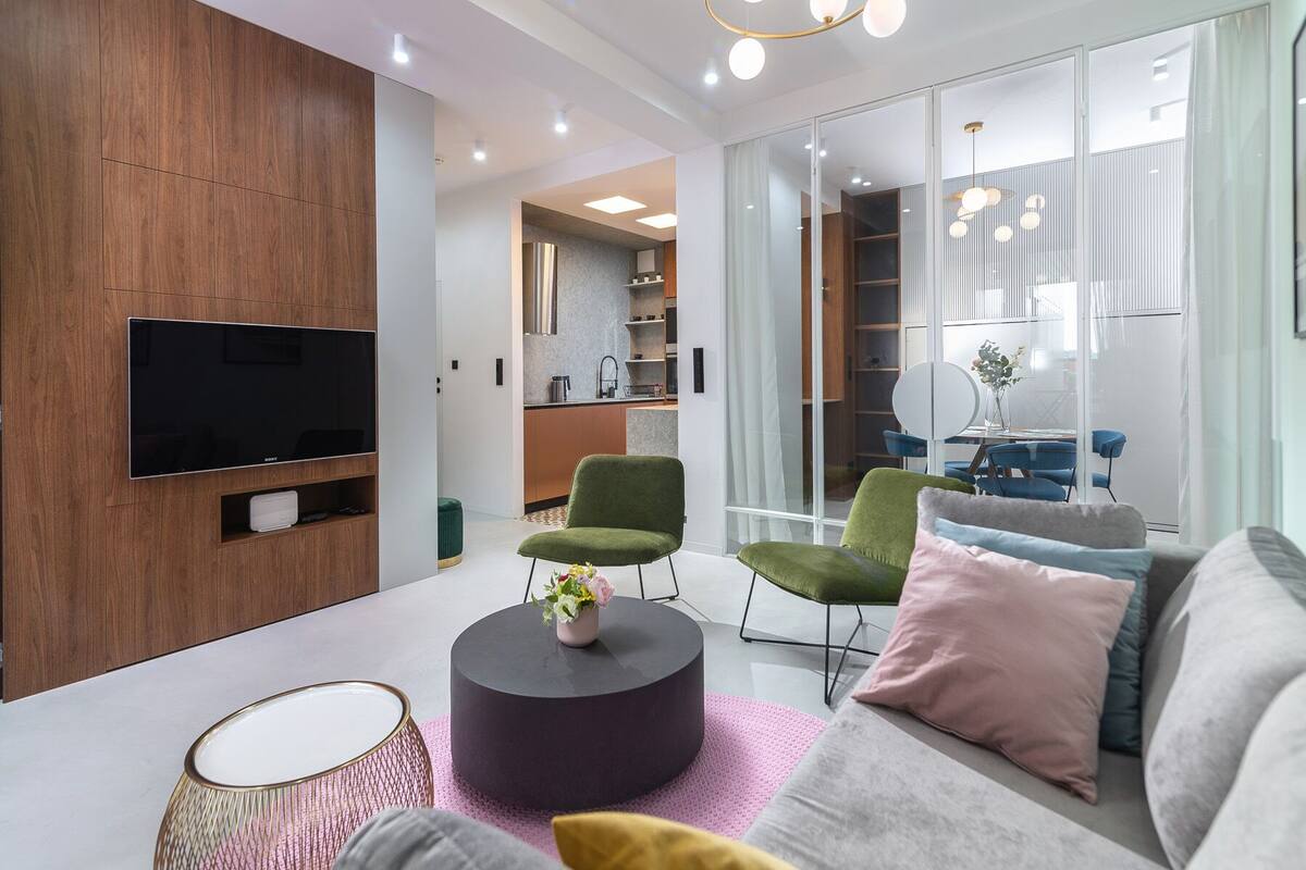 A modern living room with a flat-screen TV, green and grey chairs, a round coffee table with a vase, and a grey sofa with cushions.
