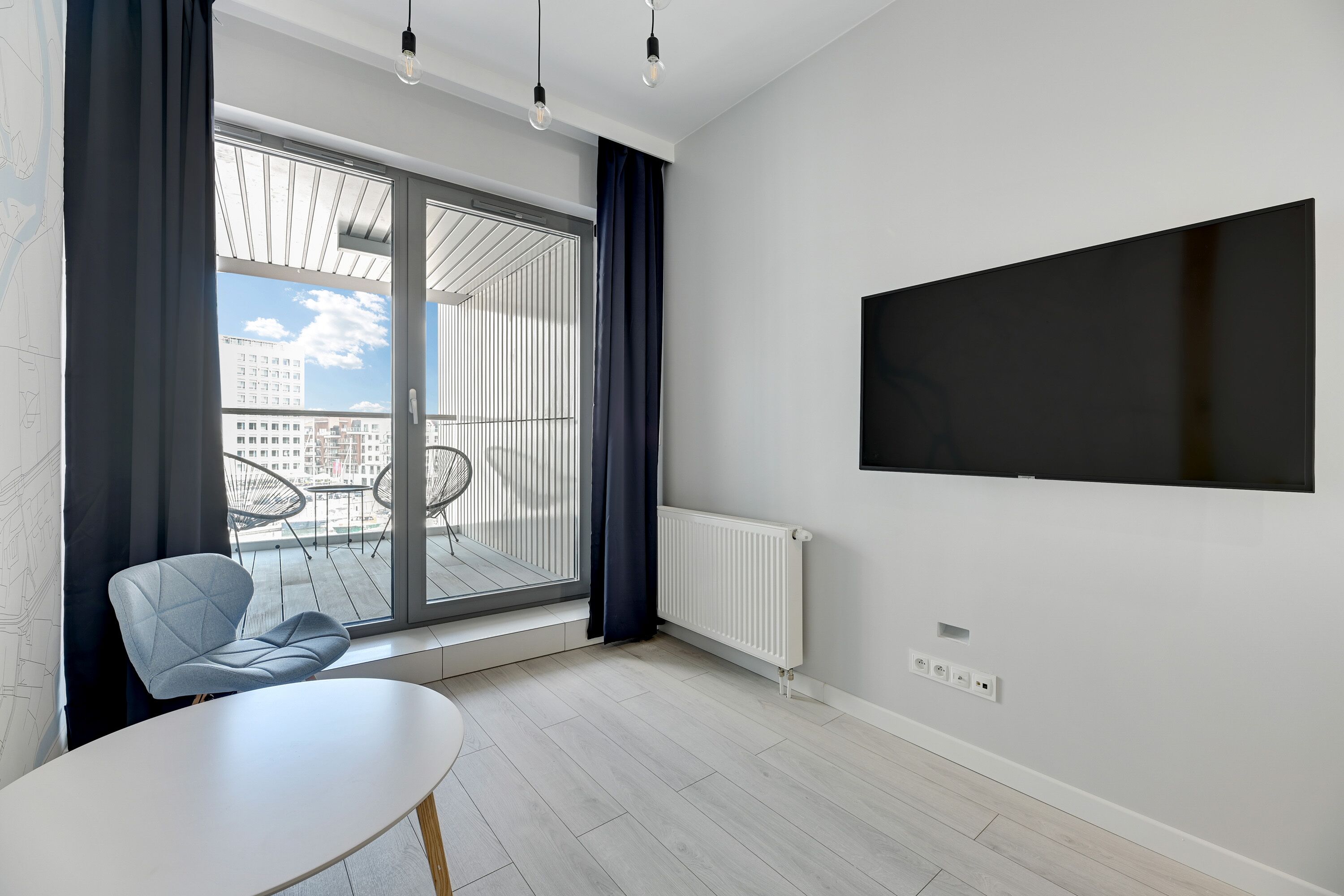 A modern living room with a large window, a flat-screen TV, a dining table, and a chair.