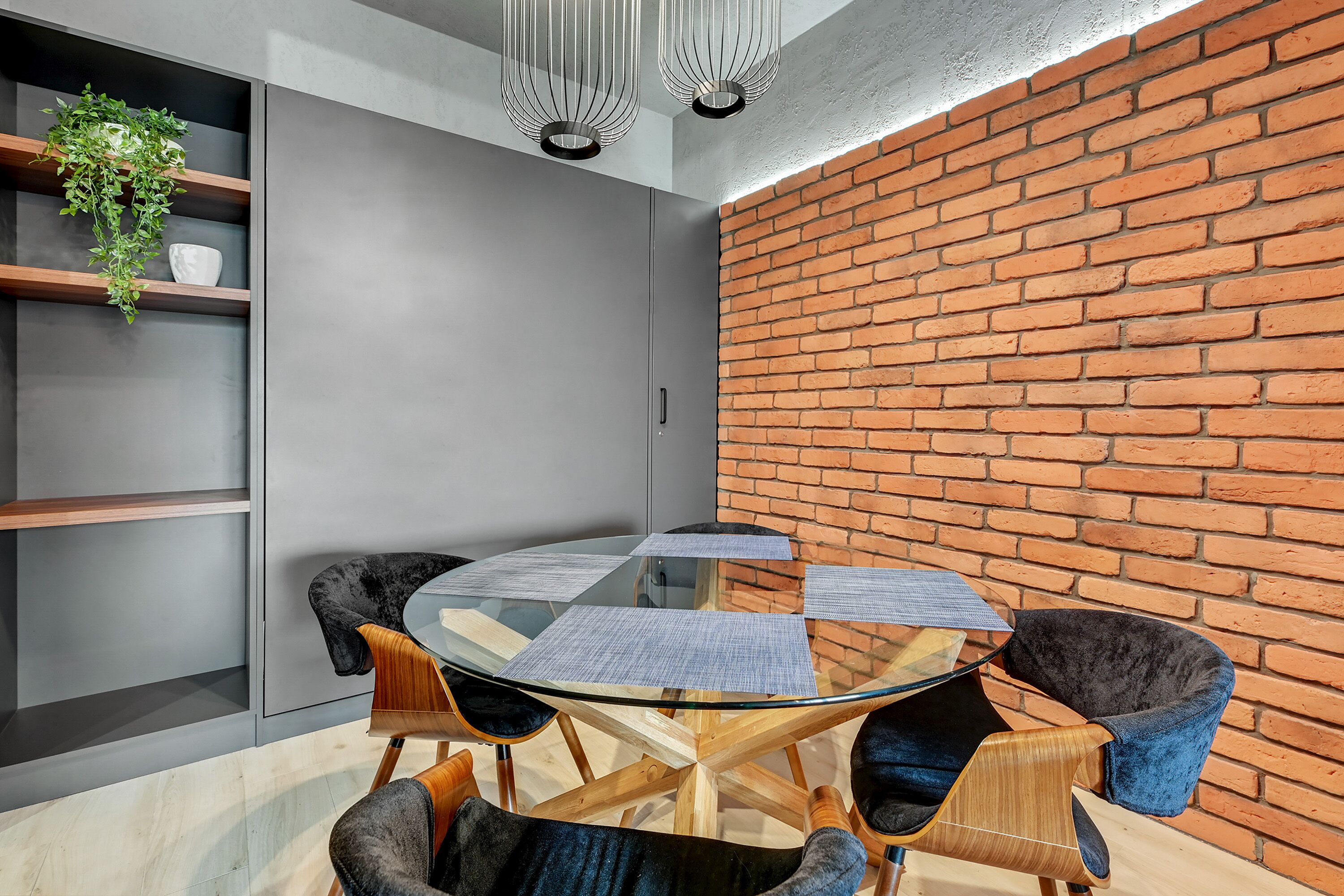 A modern meeting room with a glass table, four chairs, a brick wall, and a grey cabinet with shelves.