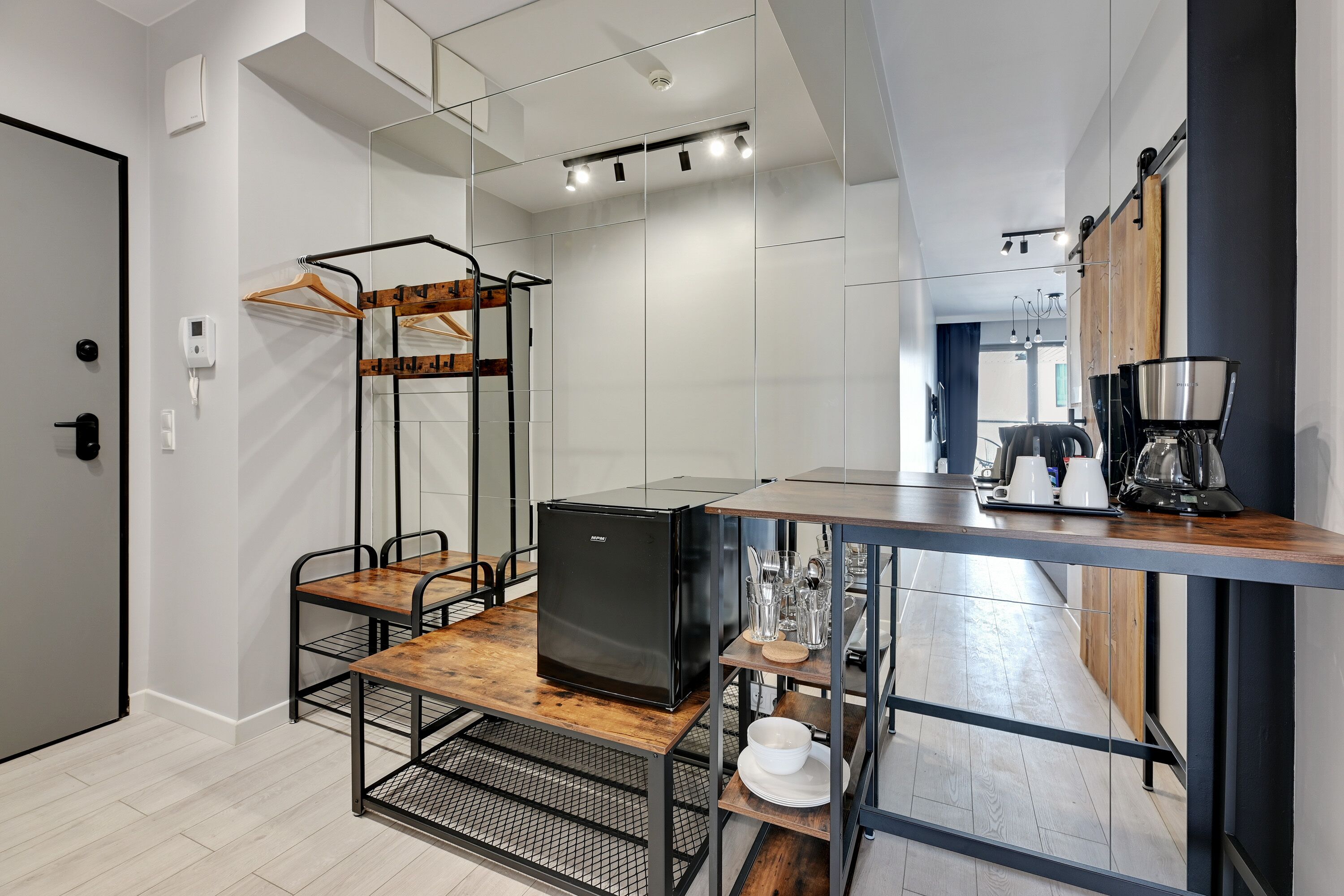 A compact kitchen with a black refrigerator, wooden table, and metal chairs. A hanging rack is mounted on the wall, and a coffee maker is on the counter.