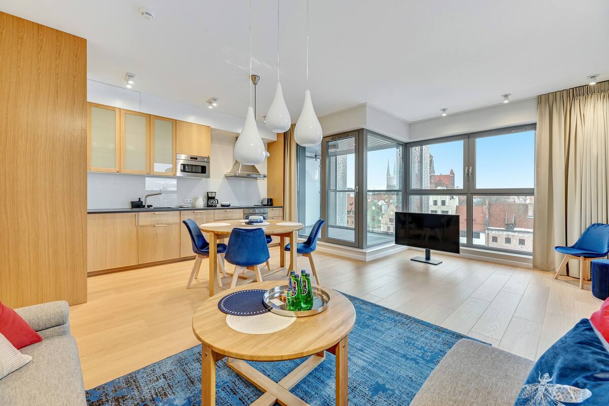 A modern apartment with a kitchen, dining area, and living room. It features large windows, a flat-screen TV, and a balcony with a city view.