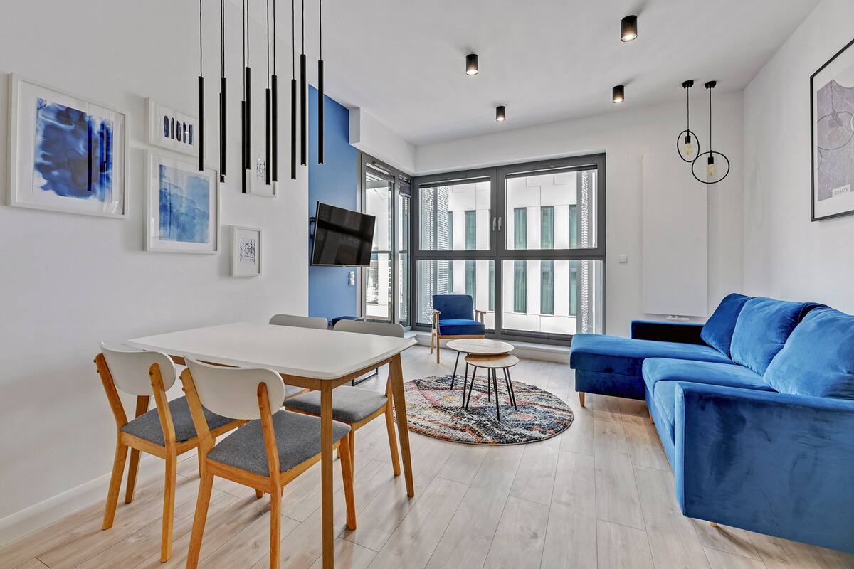A modern living room with a blue sofa, a dining table with chairs, and a TV mounted on the wall.