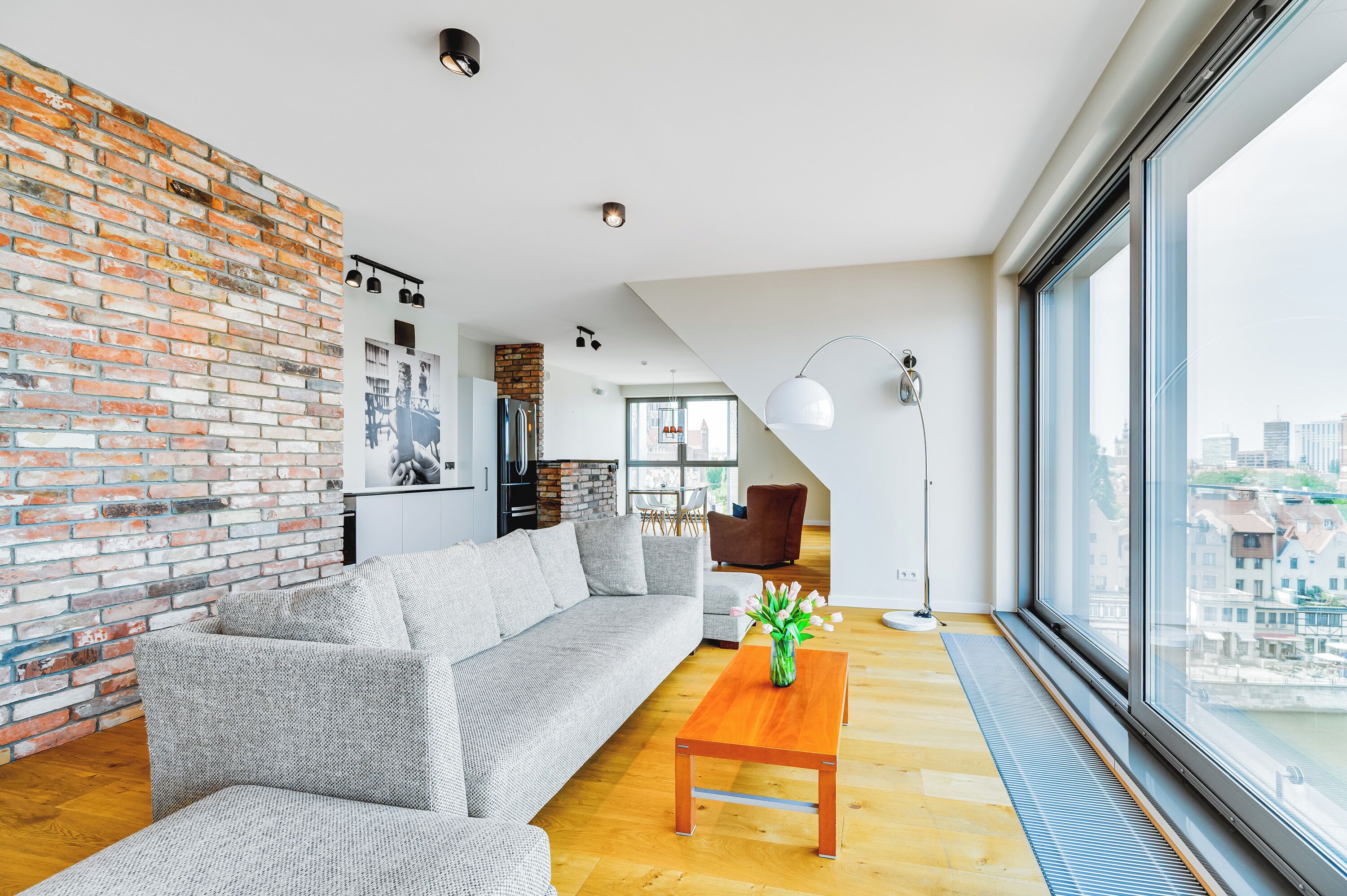 A modern living room with a brick wall, a grey sofa, a wooden coffee table, and a large window offering a city view.