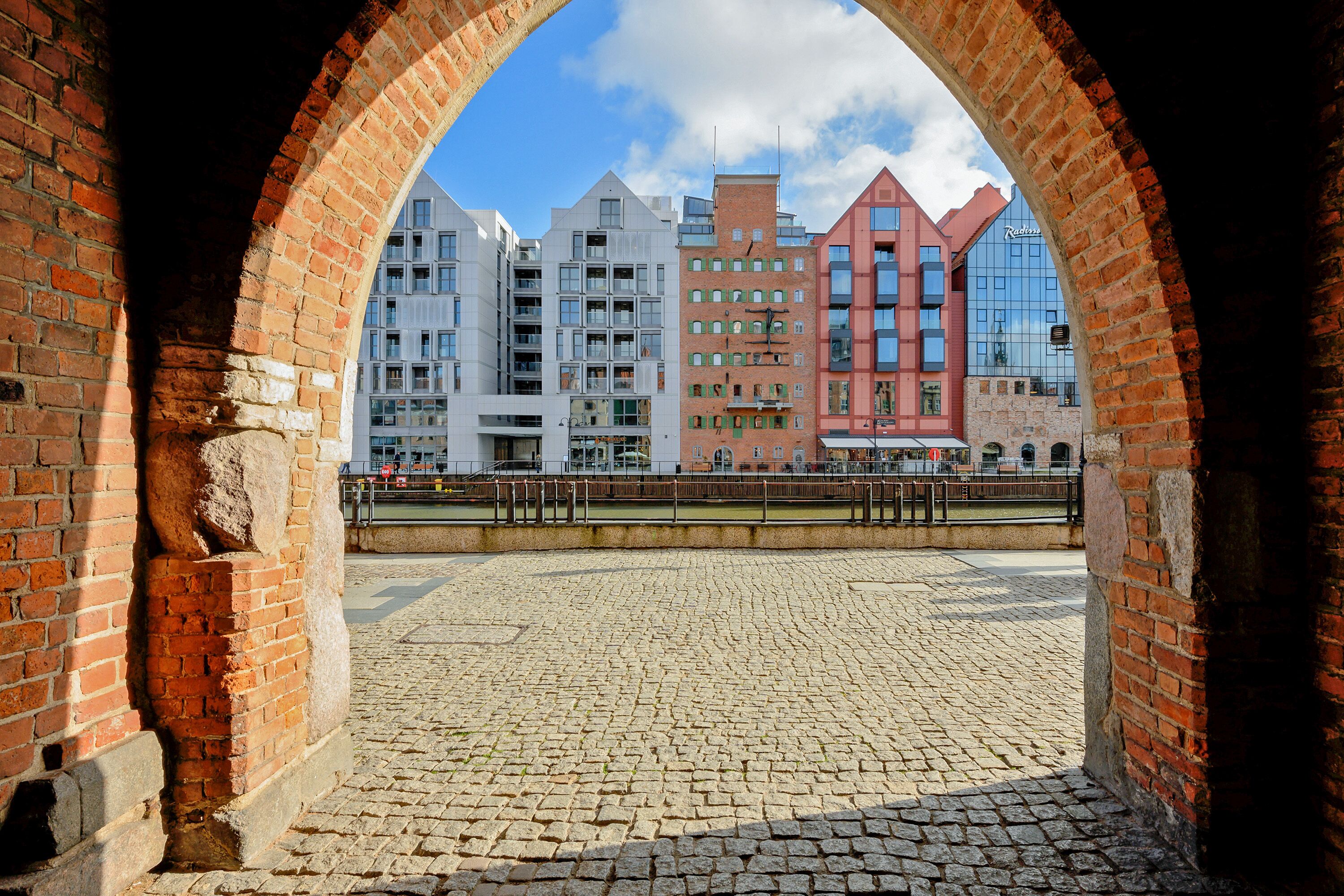 An arched brick structure with cobblestone flooring, leading to a modern urban area with contemporary buildings.