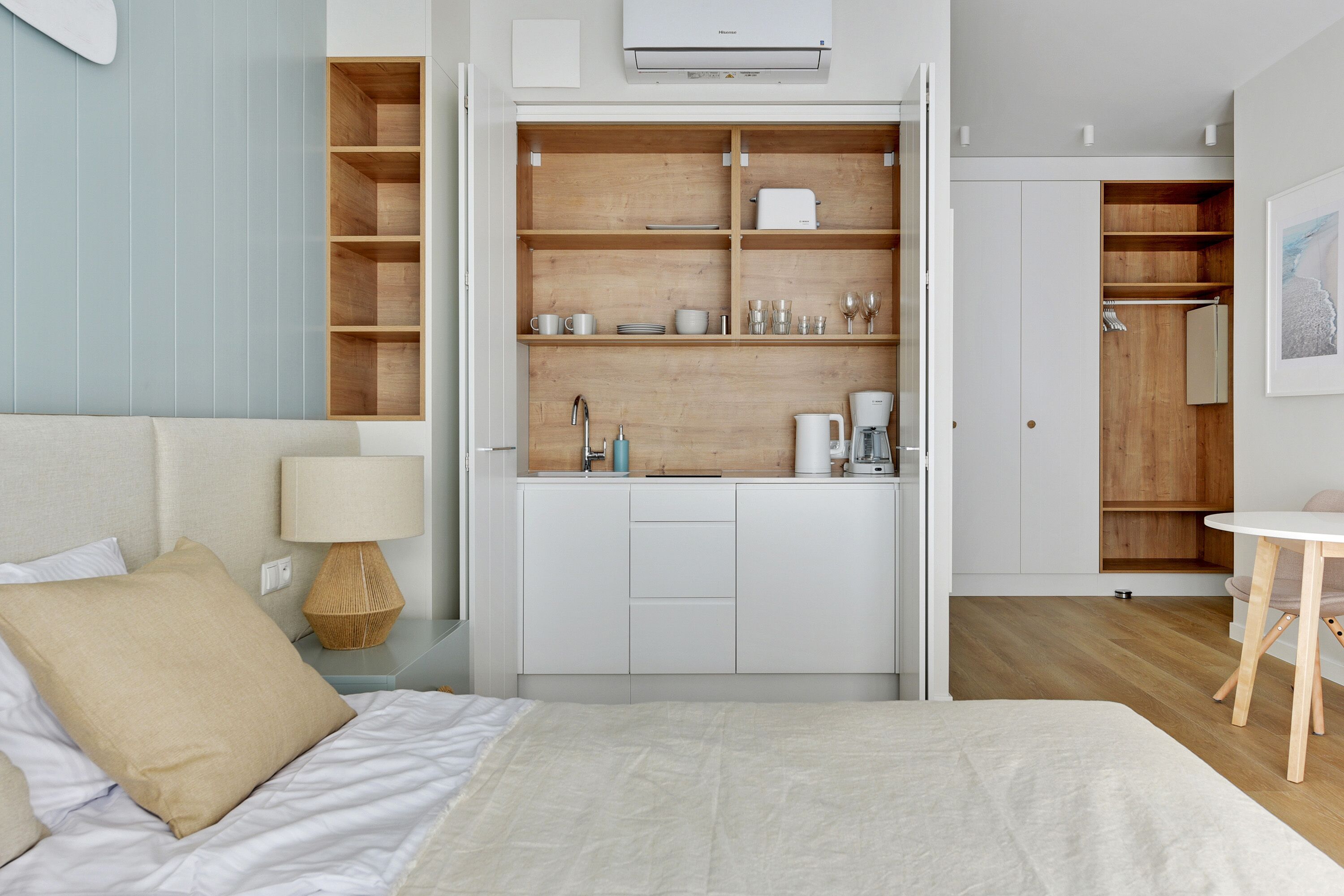 A bedroom with a bed, a bedside table with a lamp, a wardrobe with shelves, and a small table with a chair.