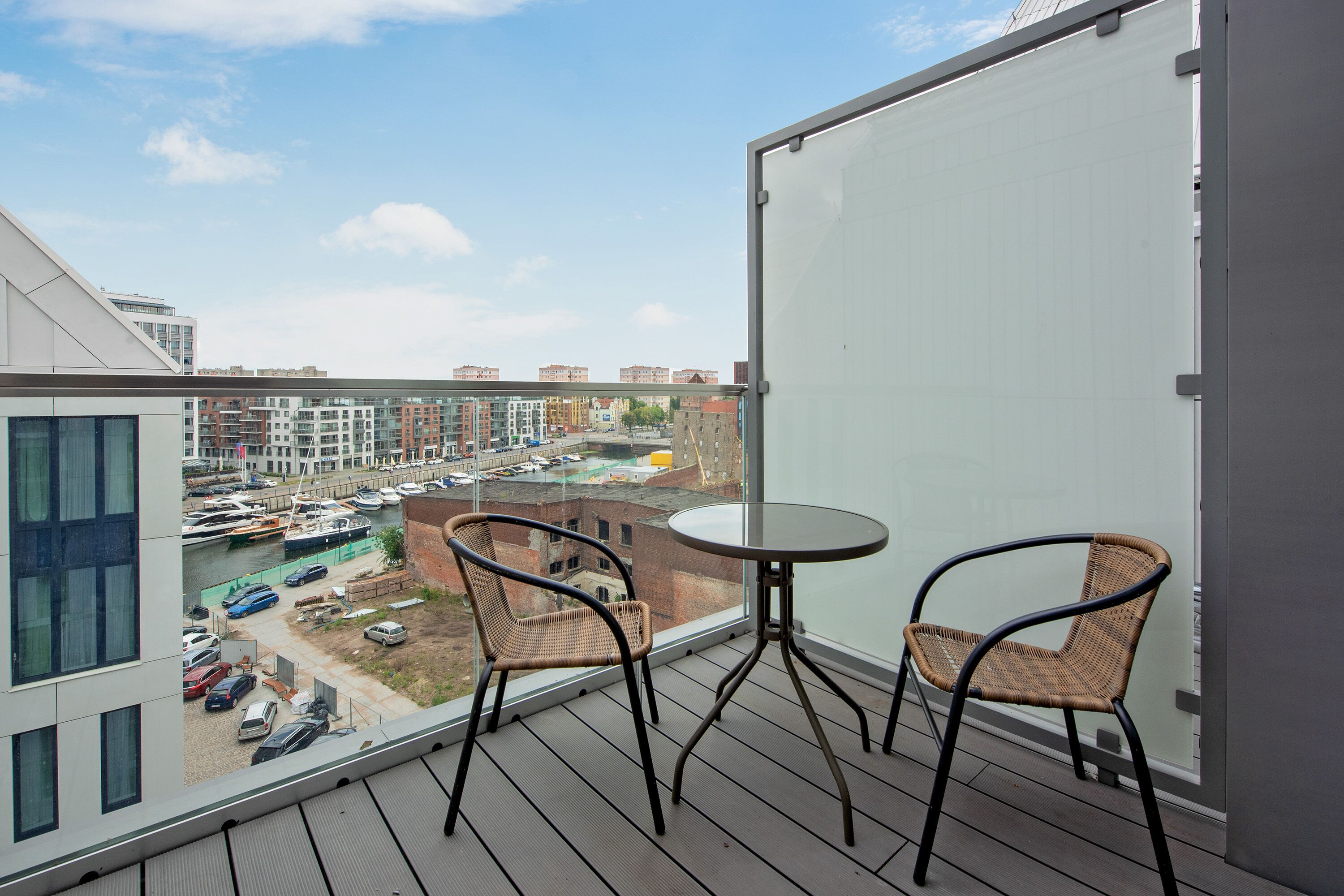 A balcony with two wicker chairs and a small table, overlooking a marina with boats and a parking lot.