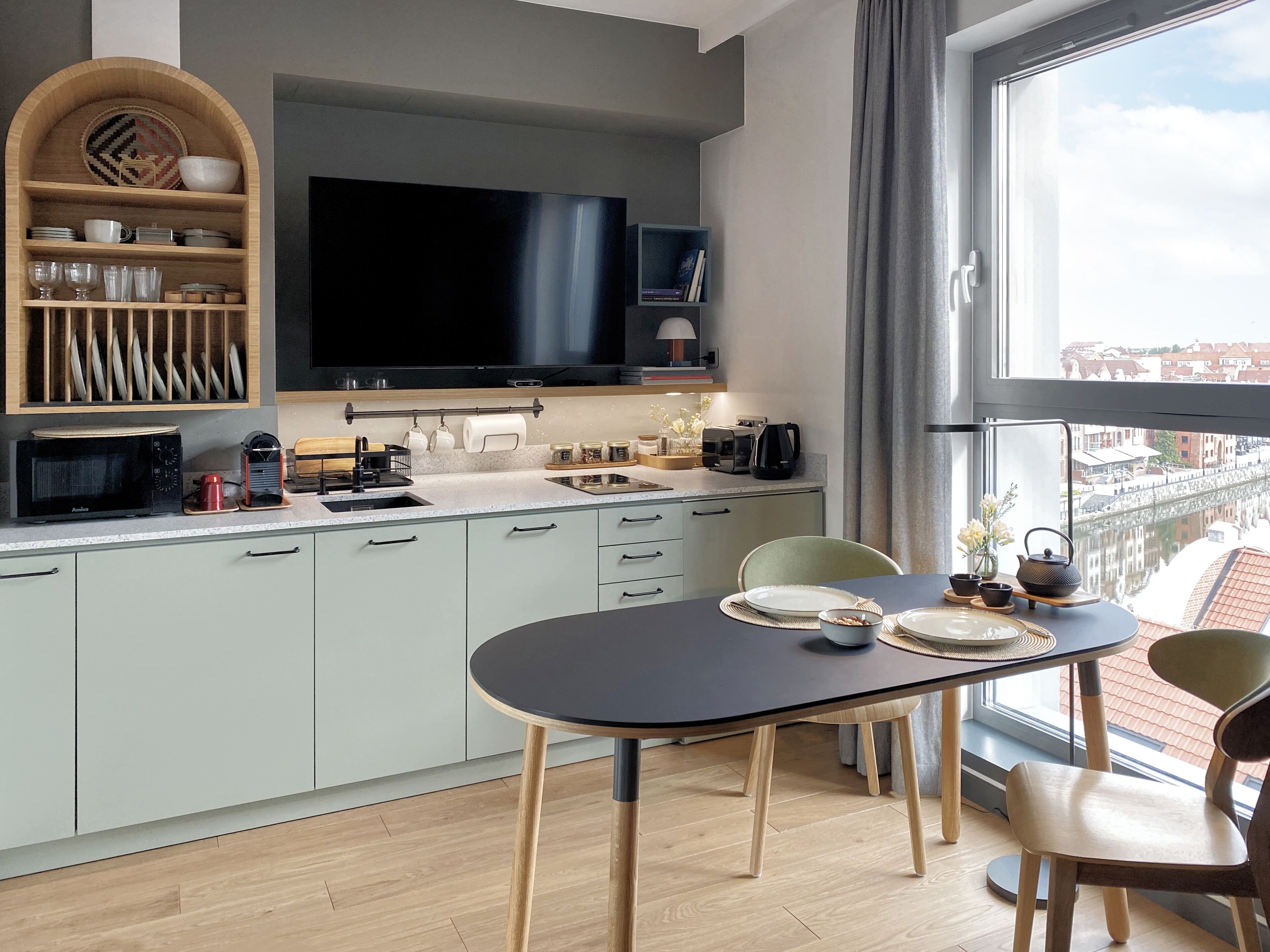 Une cuisine moderne avec un coin repas, dotée d’un téléviseur à écran plat, d’étagères en bois et offrant une vue sur un canal grâce à de grandes fenêtres.