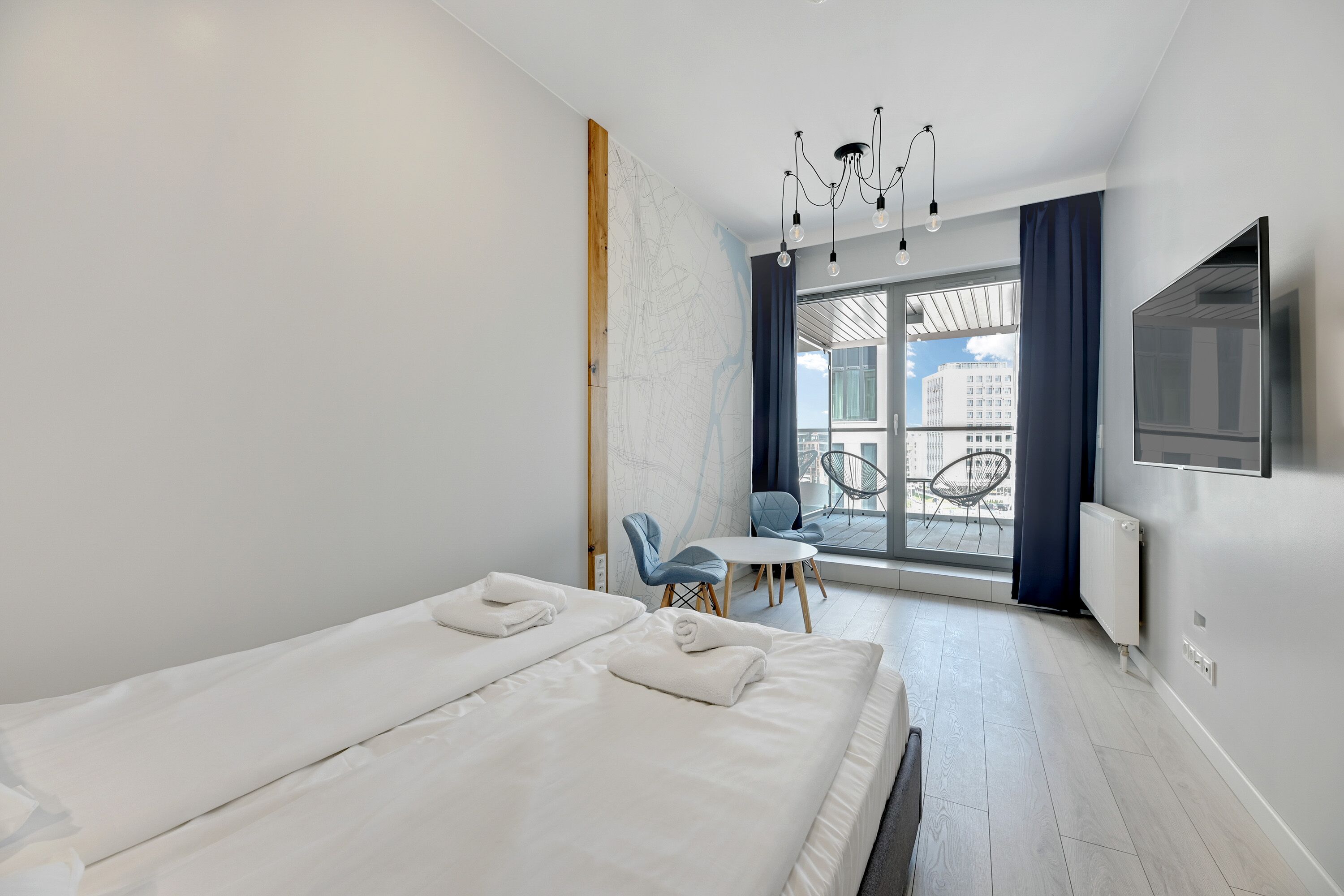Una habitación de hotel moderna con una cama grande, una mesita pequeña, dos sillas, un televisor y vistas a la ciudad a través de amplios ventanales.