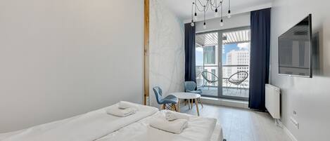 A modern hotel room with a large bed, a small table, two chairs, a TV, and a view of the city through large windows.