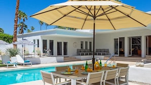 An outdoor pool area with a table and chairs, a large umbrella, and a poolside lounge with chairs and a sofa. - Apollo 1000 - Space Age Luxury Palm Springs Retreat (Palm Springs)