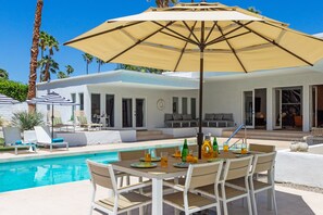 Zona de piscina al aire libre con mesa y sillas, un gran paraguas y un área de descanso junto a la piscina con sillas y un sofá.