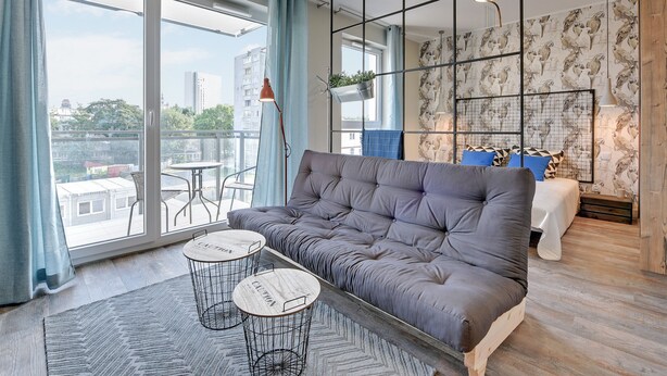 Un salon moderne avec un canapé gris, deux petites tables rondes, un tapis à motifs et une grande fenêtre avec des rideaux.