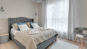 Un lit bien fait, avec une tête de lit grise, des oreillers blancs et bleus, et une couverture beige. On y trouve une table de chevet grise avec une lampe, une chaise à motifs et une grande fenêtre avec des rideaux légers.