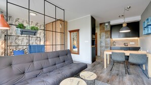 Un salon moderne avec un canapé gris, deux tables rondes et un coin cuisine doté de meubles en bois et d’un luminaire suspendu.