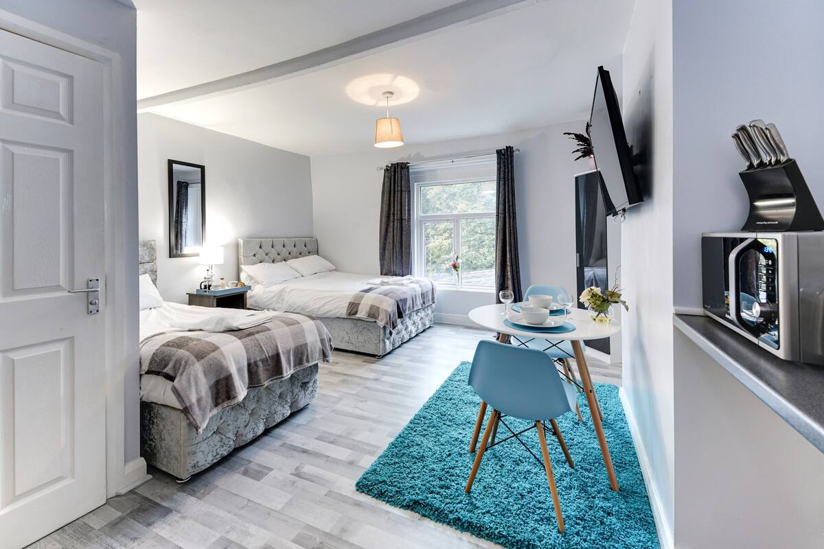 A modern bedroom with two beds, a small dining area, and a TV on the wall.