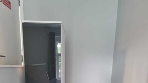 A narrow hallway with a dark floor, white walls, and a door leading to a room with a chair and a window. - Grey Stone Studio Apartments (Halifax)