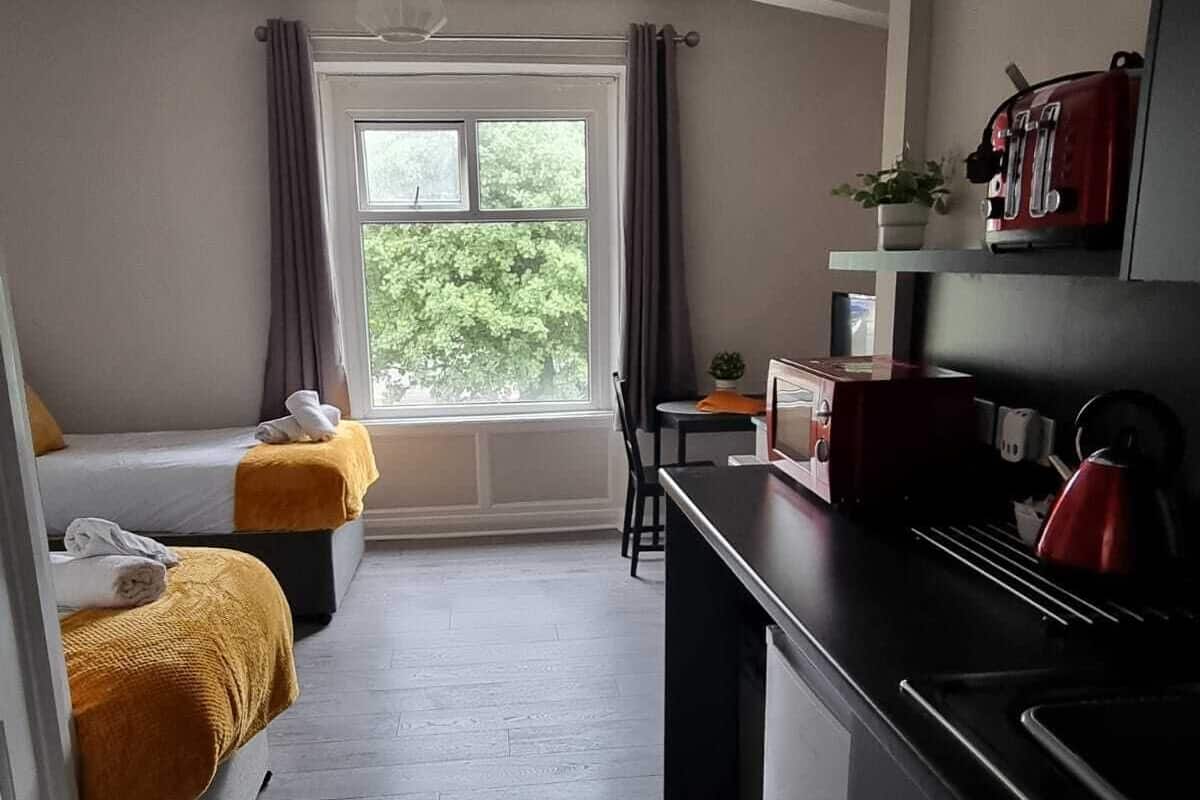 A compact hotel room with a bed, a small table, a kitchenette, and a window with curtains.
