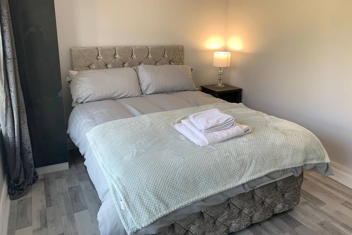 A neatly made bed with a light-colored duvet, pillows, and folded towels. There is a bedside lamp with a white shade, and the room has a simple, clean design with a wooden floor.