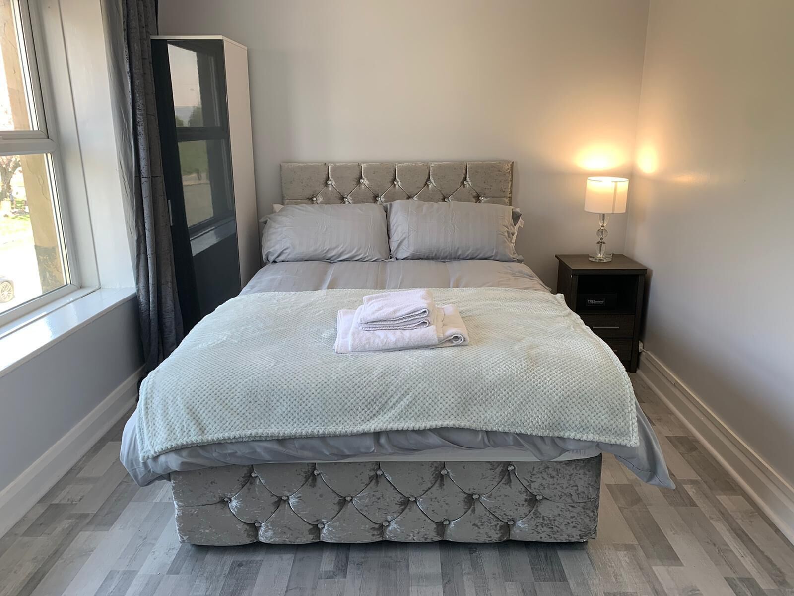 Een netjes opgemaakt bed met een gestoffeerd hoofdbord, een nachtkastje met een lamp, en een raam met gordijnen.
