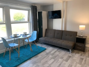 A room with a sofa, two chairs, a small table with a bowl and a cup, a TV on the wall, and a lamp on a side table. - Grey Stone Studio Apartments (Halifax)