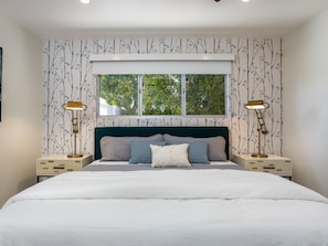 A bedroom with a large bed, two bedside tables with lamps, a window with a view of trees, and patterned wallpaper. - Fab Four at El Mirador – Midcentury Pool Retreat (Palm Springs)