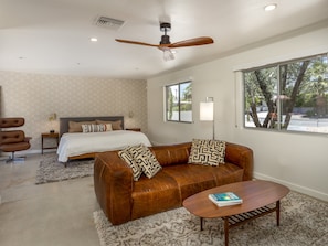 A bedroom with a bed, a brown leather sofa, a coffee table, a ceiling fan, and a window with a view of trees.
