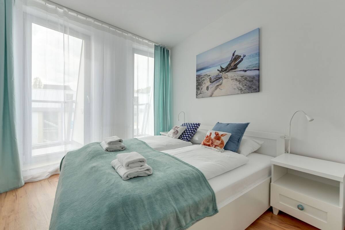 Een slaapkamer met een bed, nachtkastje, schilderij en raam met gordijnen.