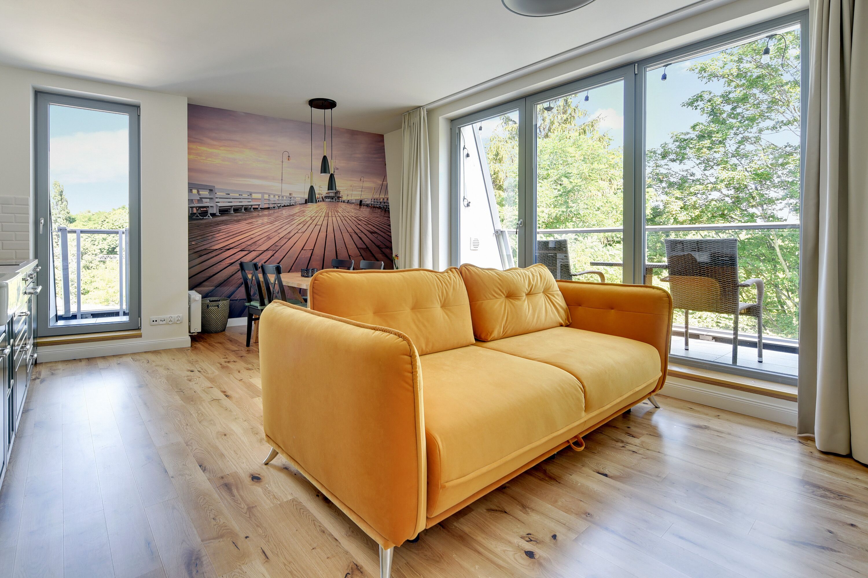 A modern living room with a large sofa, a dining area with chairs, and a mural of a pier.