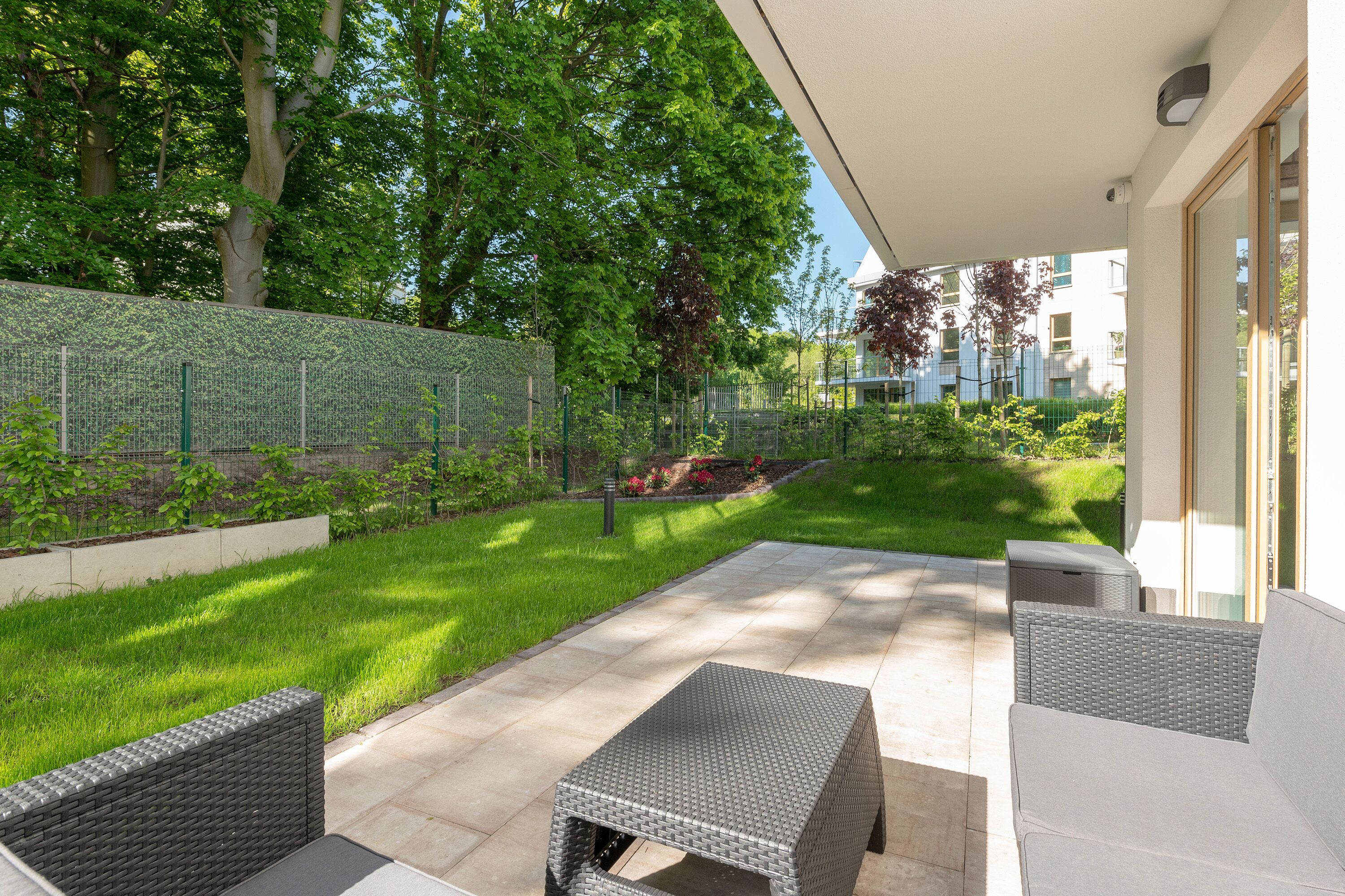 An outdoor patio with wicker furniture, a tiled floor, and a garden area with a low wall and greenery.