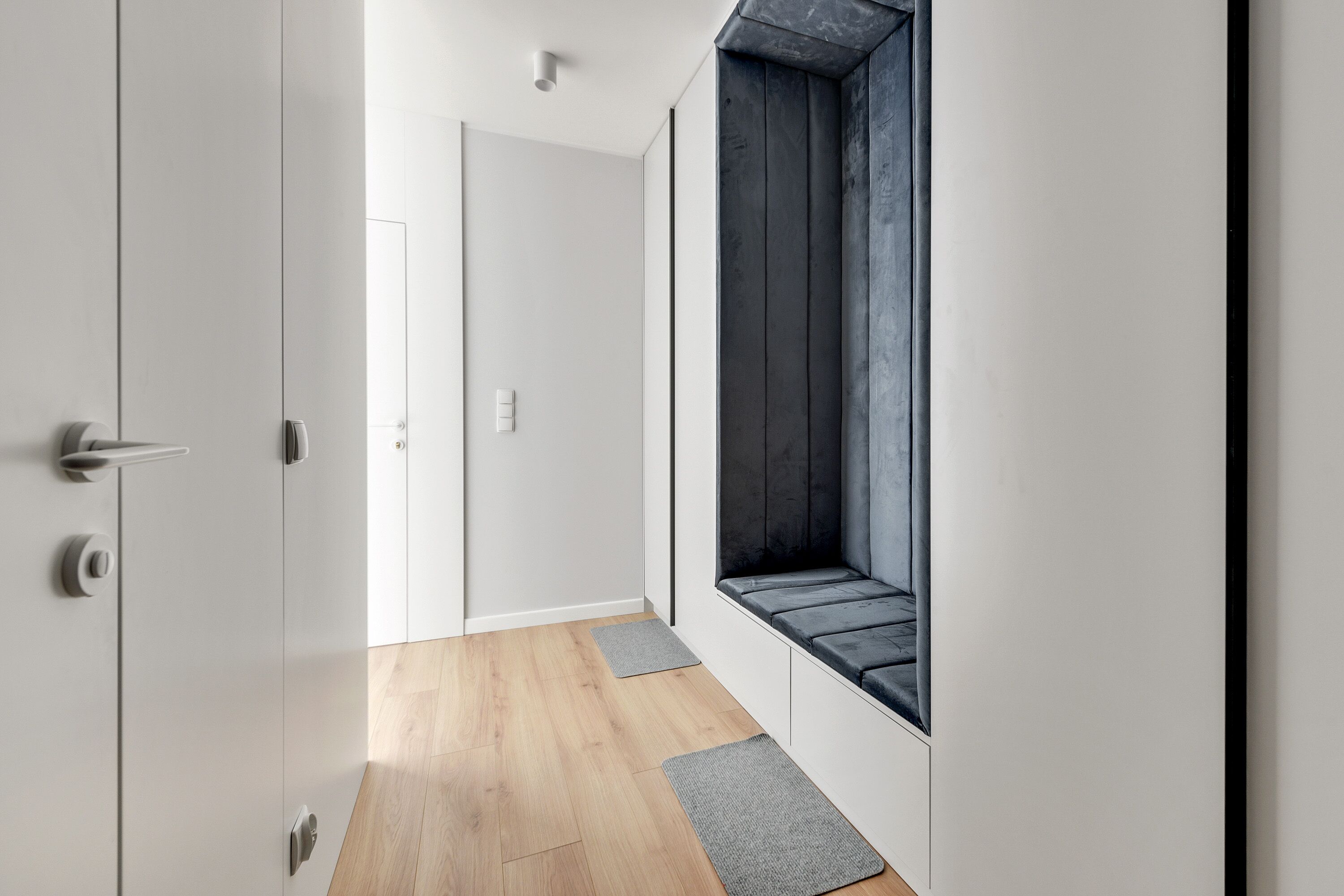 Ein moderner Flur mit Holzboden, weißen Wänden und einem großen Fenster, durch das ein dunkler Innenraum zu sehen ist.