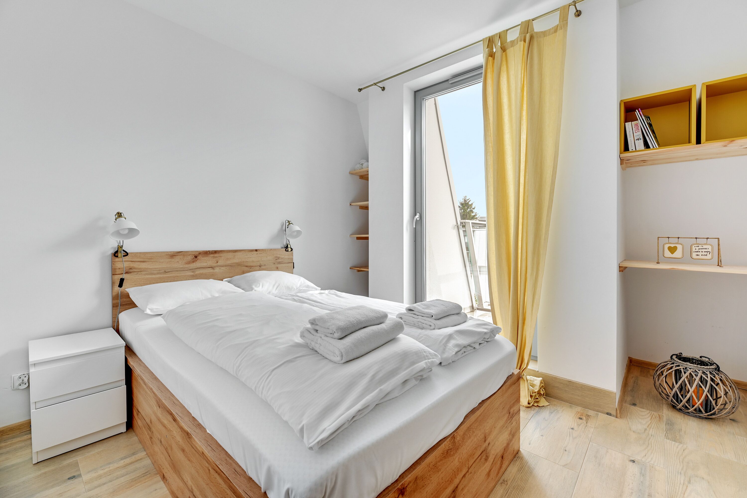A bedroom with a wooden bed, white bedding, a nightstand, a bedside table, a window with curtains, and a shelf with books and decorative items.