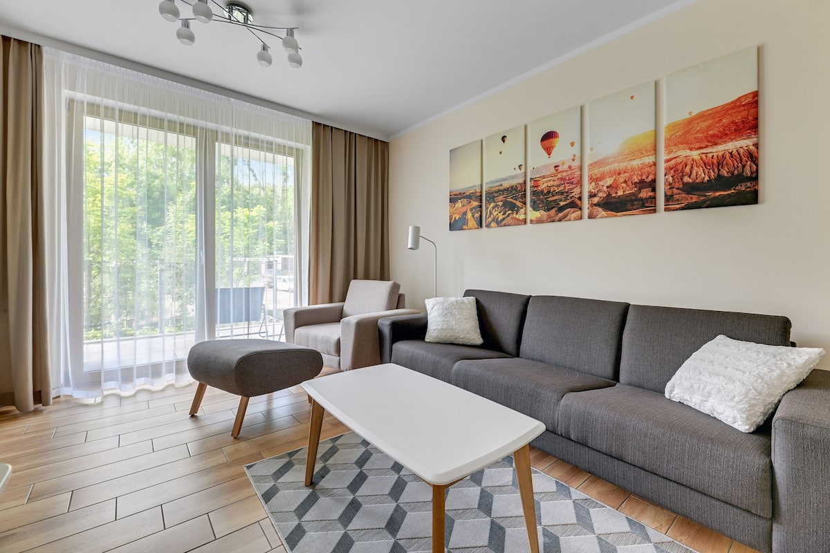 Een moderne woonkamer met een grijze bank, een witte salontafel en een groot raam met gordijnen.