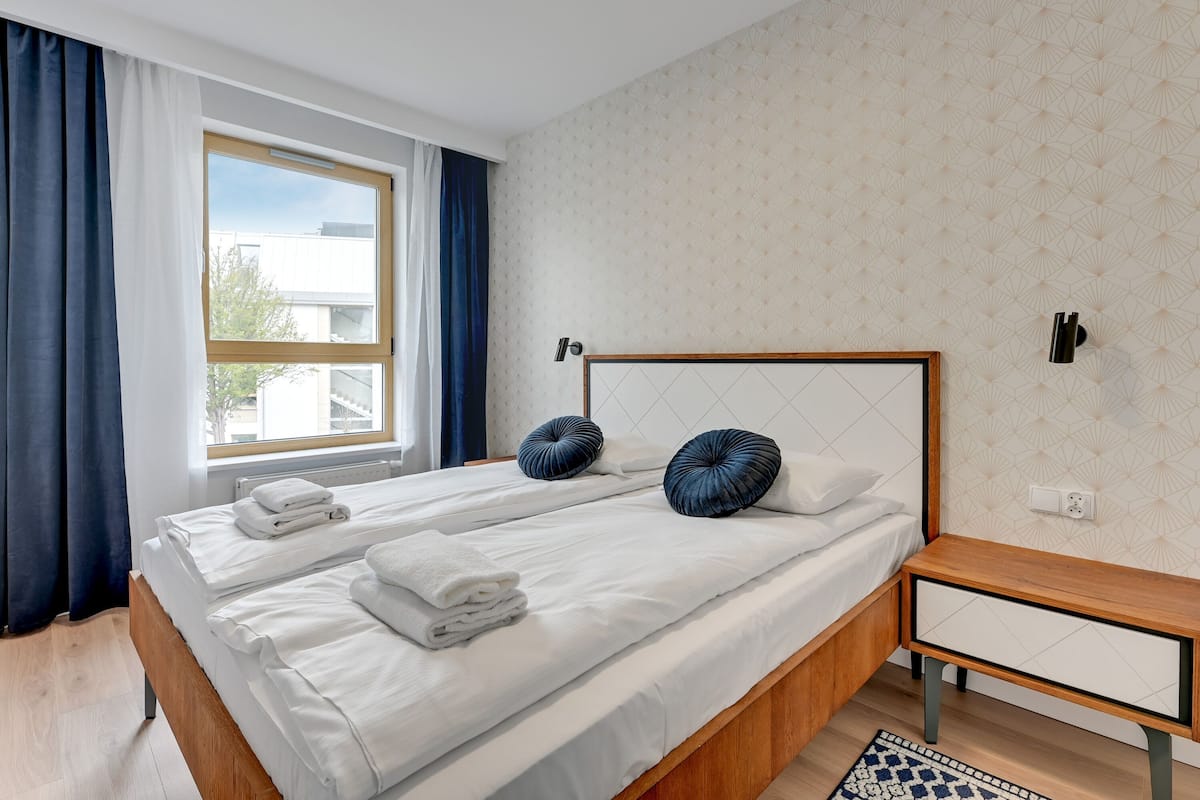 Een tweepersoonsbed met witte lakens en twee decoratieve kussens, een houten nachtkastje en uitzicht buiten door een raam met blauwe gordijnen.