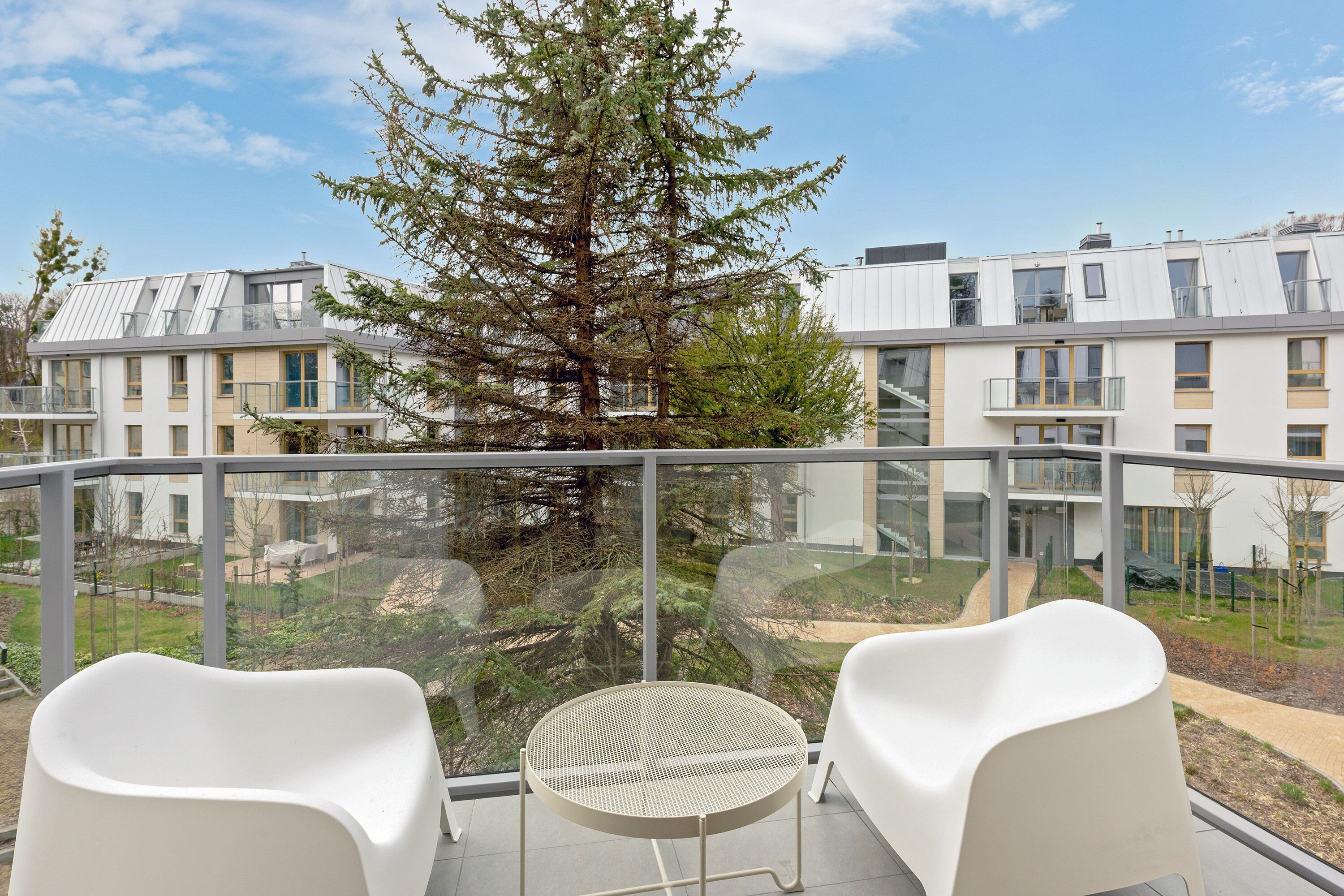 A balcony with two white chairs and a round table, overlooking a residential area with modern buildings and a large tree.