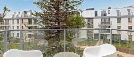 A balcony with two white chairs and a round table, overlooking a residential area with modern buildings and a large tree.