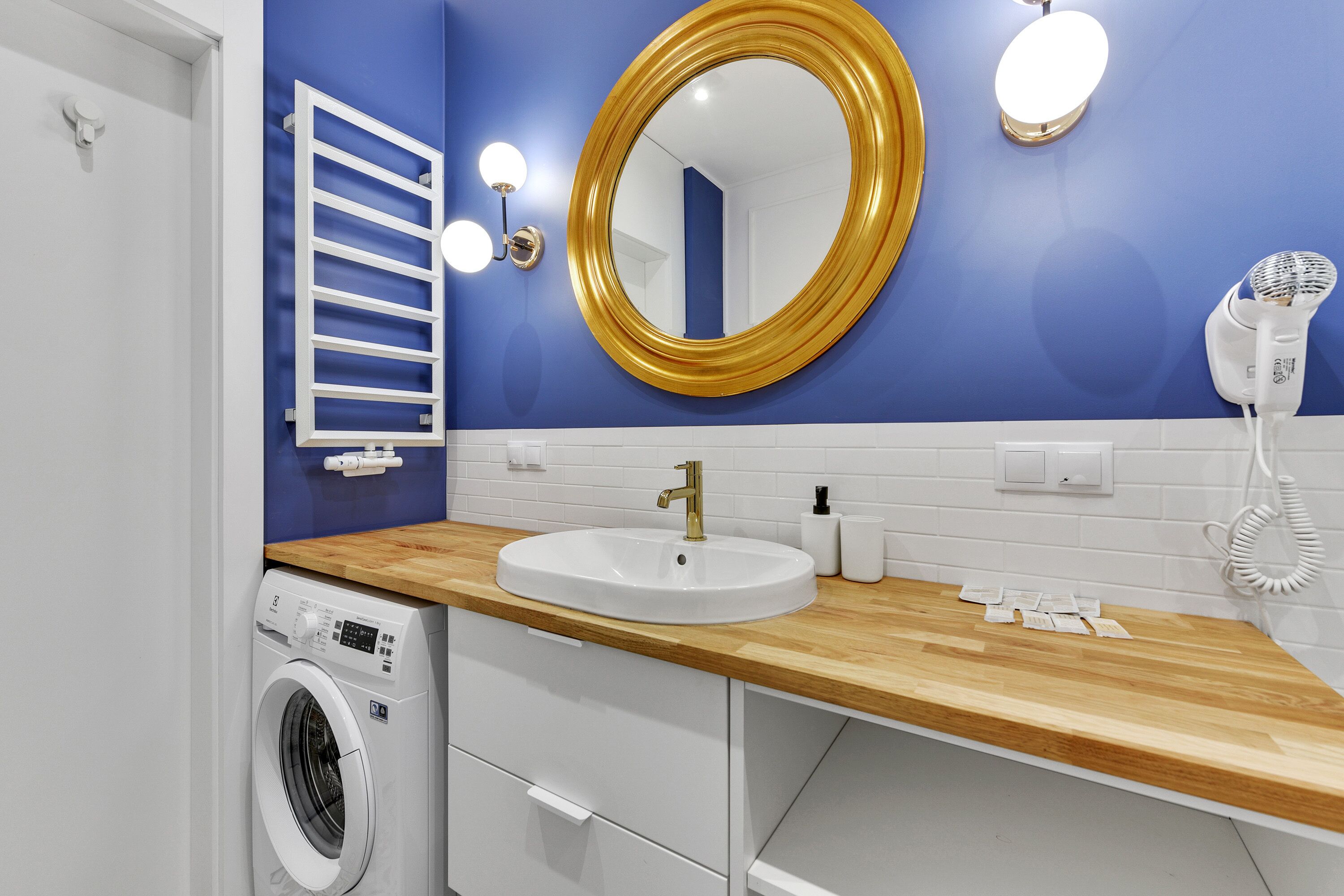 Un baño con encimera de madera, lavabo blanco, espejo con marco dorado y lavadora.